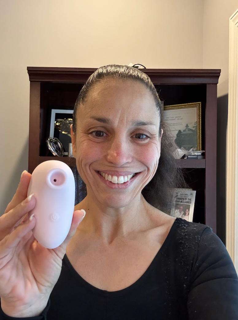 Light pink skincare device held by a woman in a black top, set against a home office background.
