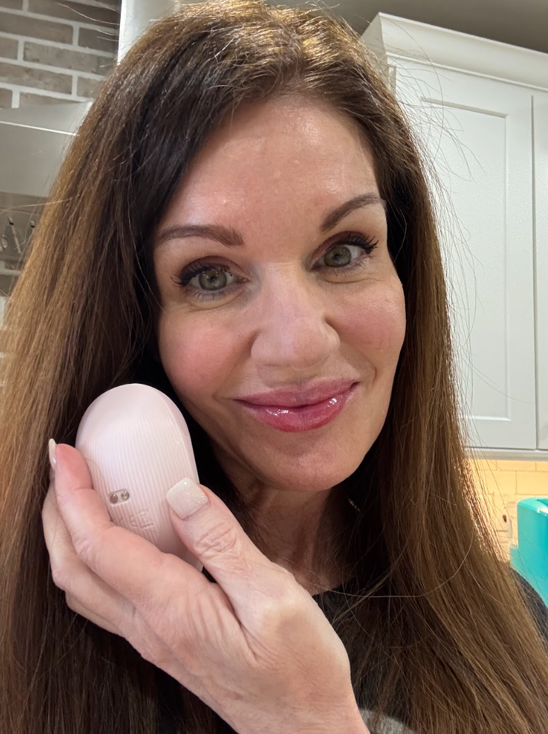 Light pink facial cleansing device held by a woman with long brown hair, smiling in a kitchen setting.