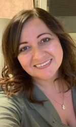 Smiling woman with wavy brown hair wearing a green top and a pendant necklace.