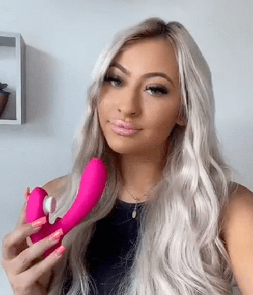 Pink dual-action vibrator with ergonomic design, held by a woman with long, wavy blonde hair.