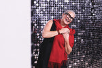 Smiling woman in black dress and oversized glasses, wearing a red scarf, against a sequin backdrop.
